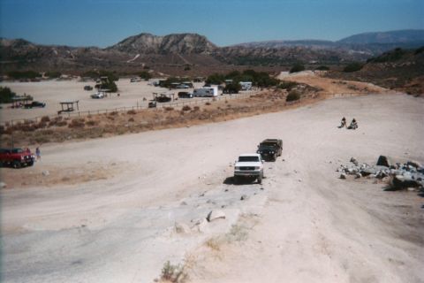 1990 Ford Bronco - white truck and a dark Hummer travel a wide sandy trail at a desert OHV camp with motorcycles and RVs in view.