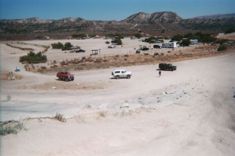1990 Ford Bronco - elevated view of a desert OHV staging area shows red, white, and dark trucks on sand among scattered RVs.