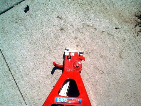 1995 Ford Bronco - A red hydraulic jack photographed from above along its arm on a sunlit concrete driveway, with a toothed lift rack and a blue-and-white 'HR…