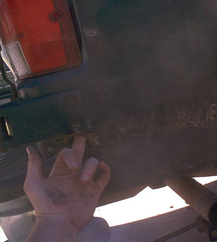 1995 Ford Bronco - Dim, dusty shot under the rear corner of a truck or SUV, with the red tail light lens and body-mount bolts visible at upper left and a hand…