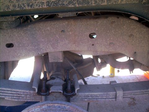 1995 Ford Bronco - Under-vehicle view looking up at a rusty rear leaf spring pack clamped to the axle with long U-bolts and a spring plate, framed by the fram…
