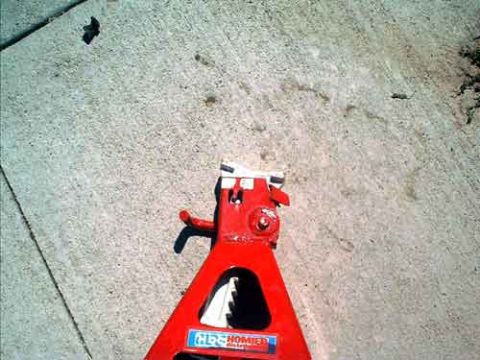 1995 Ford Bronco - Overhead shot of a red steel jack stand standing on a concrete driveway, showing its ratcheting toothed post, saddle and release lever. A b…