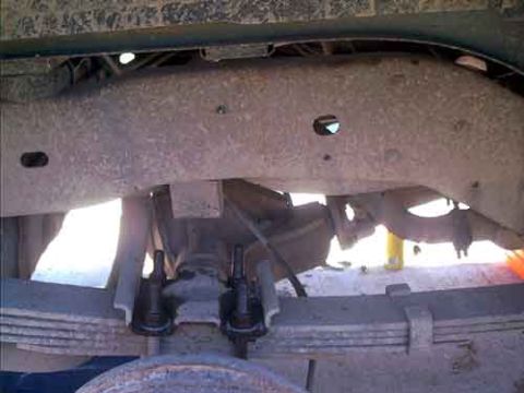 1995 Ford Bronco - Under-vehicle view of a solid front axle and leaf-spring suspension, looking up at the frame crossmember with the tie rod and drag link end…