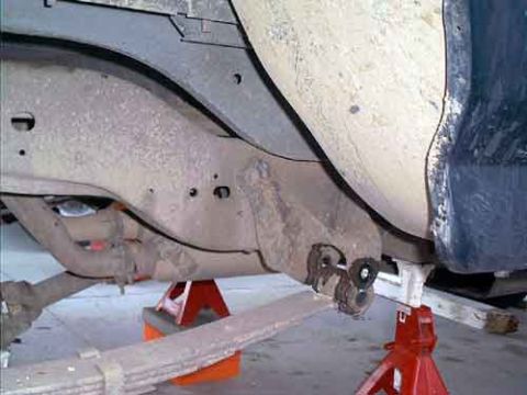 1995 Ford Bronco - Underside view of a truck raised on red jack stands during a lift install, showing the dirty frame rail, body mount bracket, and exhaust/ca…