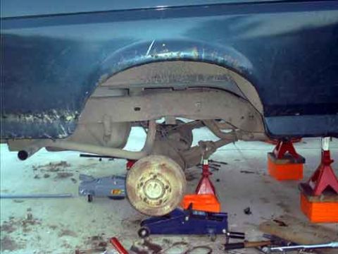 1995 Ford Bronco - The rear of a dark blue vehicle raised on orange jack stands in a garage with a wheel removed, exposing the rear axle and brake hub; a floo…
