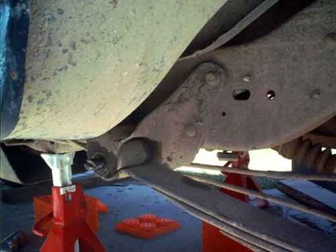 1995 Ford Bronco - Underside of a truck or SUV supported on red jack stands, viewed from below to show the frame rail, a leaf-spring, and axle components coat…
