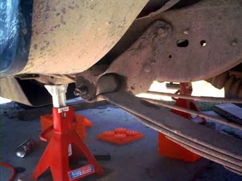 1995 Ford Bronco - Undercarriage of a truck or SUV raised on red jack stands, showing a rusty frame rail, leaf-spring pack, and axle components caked with dri…