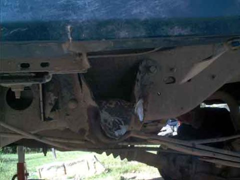 1995 Ford Bronco - View underneath a blue vehicle raised on a jack stand over grass, showing the dirty undercarriage, floor pan, frame crossmember and axle ho…