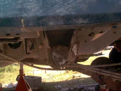 1995 Ford Bronco - Underside view of a blue truck raised on red jack stands in a grassy yard, showing a dusty frame crossmember and stripped-out front suspens…