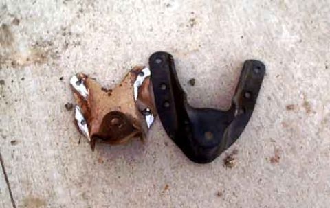 1995 Ford Bronco - Two removed suspension mounting brackets lying side by side on concrete: a badly rusted, torn-through bracket next to an intact black V-sha…