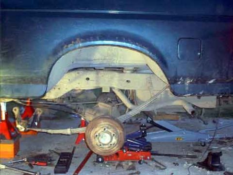 1995 Ford Bronco - Underside rear view of a dark blue Ford Bronco raised on a red floor jack, showing the rear wheel, leaf spring suspension, differential, an…