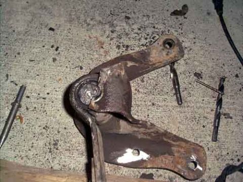 1995 Ford Bronco - Rusty steel suspension bracket with a bushing eye, removed and lying on a concrete floor next to drill bits and punches during a suspension…
