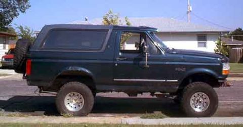1995 Ford Bronco - A dark green two-door Ford Bronco SUV with a matching hardtop shown in side profile, lifted on chunky off-road tires with white/silver whee…