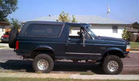 1995 Ford Bronco - Dark blue-green full-size Ford Bronco photographed from the driver's side at the curb, lifted on oversized mud tires and white-spoke wheels…