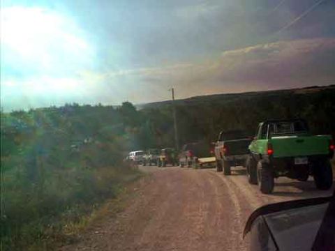 1995 Ford Bronco - A line of trucks, including a green 1995 Ford Bronco, drive down a dirt road on a sunny day.