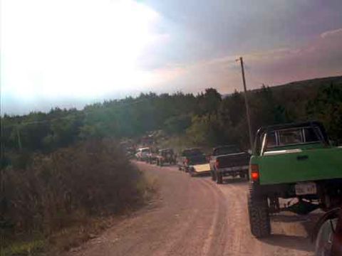 1995 Ford Bronco - A convoy of off-road vehicles travels down a dusty dirt road through a wooded area.