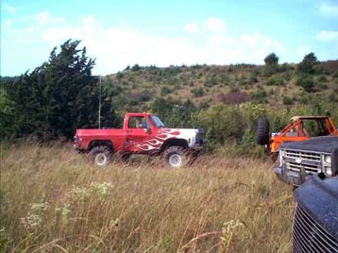 1995 Ford Bronco - Driver-side view of a lifted red-and-white flame-painted square-body pickup with the top removed, sitting on oversized mud tires in a field…