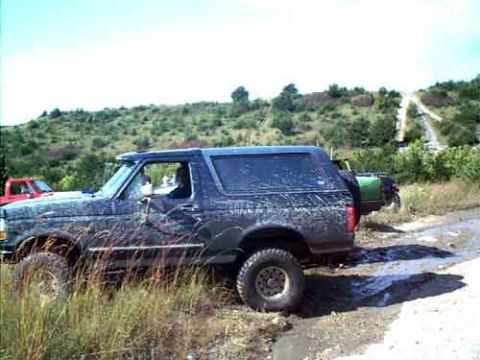 1995 Ford Bronco - Side view of a dark Ford Bronco caked in mud spray, sitting on a wet, rutted dirt trail at an off-road park. It has a hardtop, roof rack an…