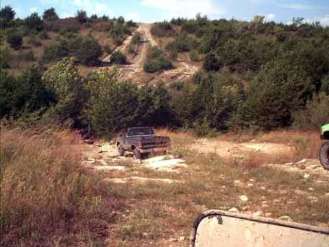 1995 Ford Bronco - A pickup truck is parked on a dirt path, with a steep, wooded hill and a dirt road leading up it in the background.