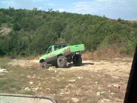 1995 Ford Bronco - A lime-green lifted mini-truck with a stripped bed and oversized mud tires climbs a rocky dirt bank at an off-road park, viewed from the re…