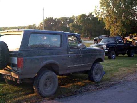 1995 Ford Bronco - Rear three-quarter view of a mud-splattered dark Ford Bronco with its hardtop on, sitting on black steel wheels and mud tires on a grassy l…
