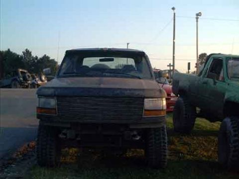 1995 Ford Bronco - A dark Ford Bronco is parked next to a green pickup truck under a clear sky.