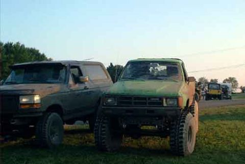 1995 Ford Bronco - Two mud-caked 4x4s parked side by side on grass at dusk: a lifted full-size Ford Bronco on the left and a green compact pickup with a chopp…