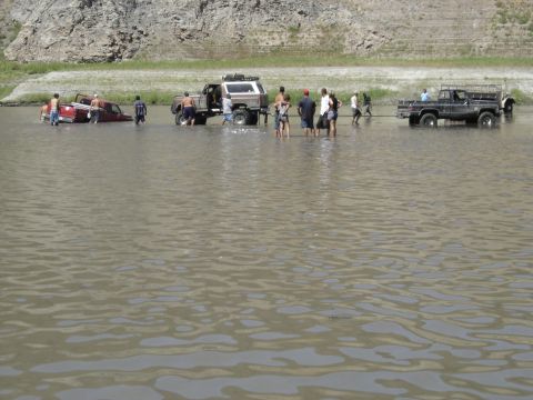 1990 Ford Bronco - crowd wades in shallow muddy water among several pickup trucks during an off-road gathering.