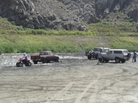 1990 Ford Bronco - ATV, a pickup towing a trailer, and SUVs gather on a gravel riverbed beside rocky bluffs.