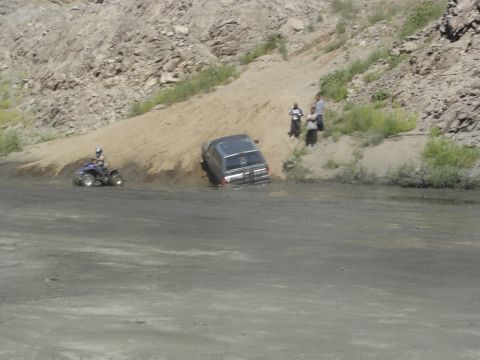 1990 Ford Bronco - ATV and a dark SUV work along a steep sandy embankment beside water while two people watch.