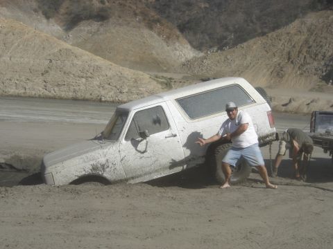 1990 Ford Bronco - man poses beside a mud-caked white Ford Bronco bogged down in a sandy pit as another person hooks a chain.