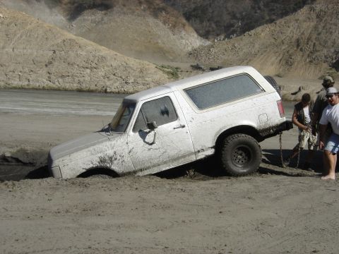 1990 Ford Bronco - mud-spattered white Ford Bronco sits tilted and stuck in a sandy pit while people attach a recovery chain.