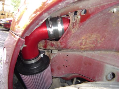 1998 Ford Taurus - Close-up of a red vehicle's fender and engine bay, with a visible aftermarket air intake and rust. Another vehicle is in the background.