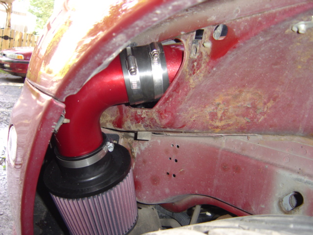 Red vehicle fender with rust, showing a red aftermarket air intake system. Another red vehicle is visible in the background