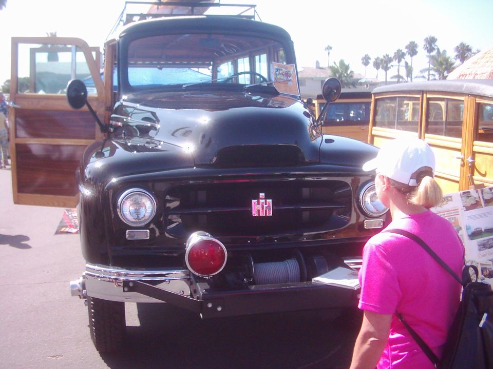 Front view of a black International Harvester woodie truck with doors open, a woman in a pink shirt looks at a display next to it. Another woodie vehicle is parked behind
