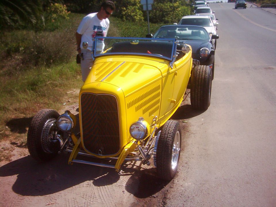 Yellow hot rod roadster with exposed engine and wide rear tires parked on dirt road