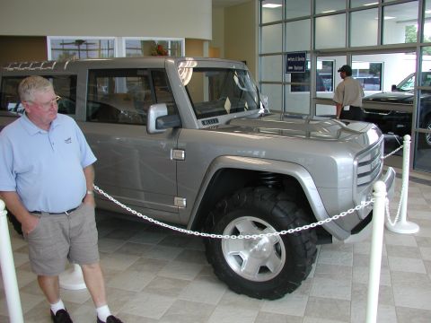 1985 Ford Bronco - A man stands beside a silver concept SUV on display in a dealership showroom.