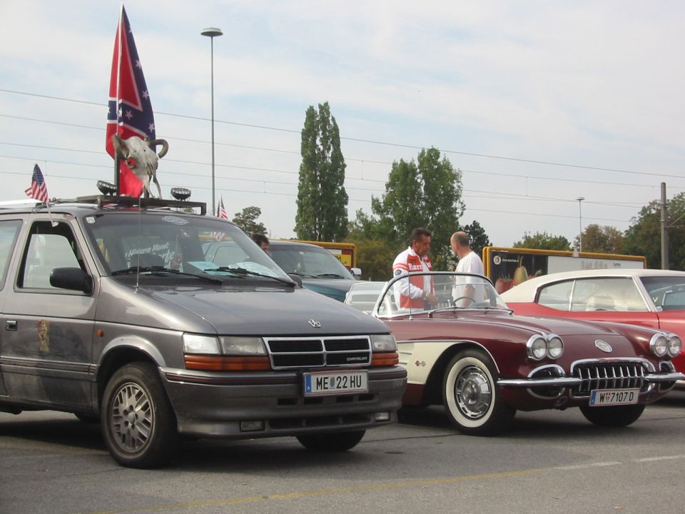 Gray minivan with Confederate flag and bull skull on roof, parked next to a red classic convertible and a red classic coupe