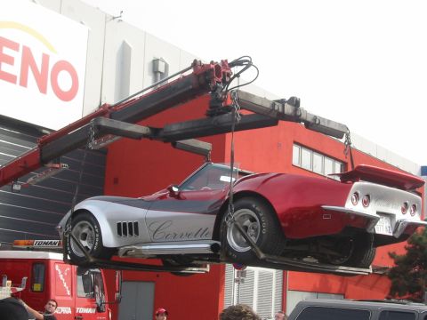 1992 Ford Crown Victoria - A red and silver Corvette is hoisted by a crane in front of a red building, with a tow truck visible below.