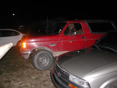 1985 Ford Bronco - A red SUV is parked closely between two other vehicles at night.