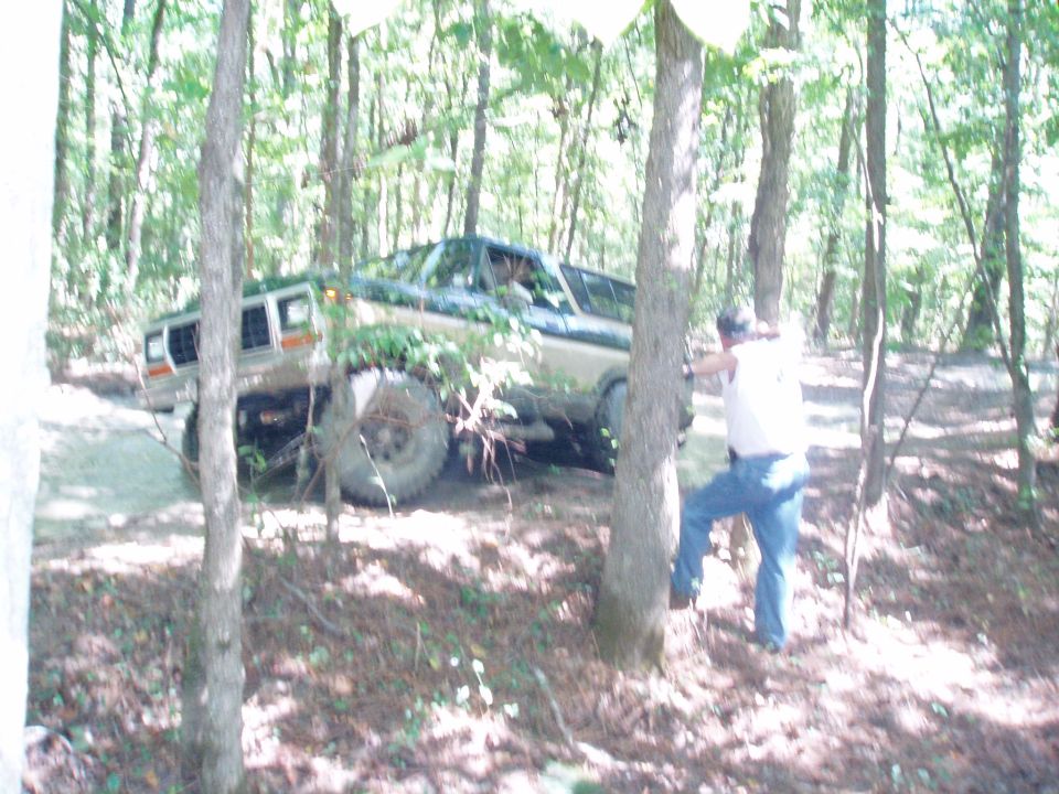 Lifted tan and green Ford Bronco on big tires driving a wooded off-road trail as a man watches