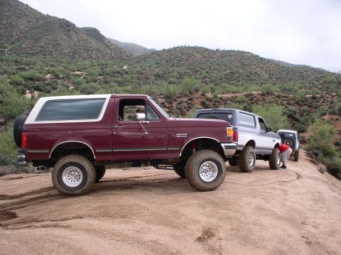 1990 Ford Bronco - Three Ford Bronco SUVs are parked on a dusty trail in a hilly, arid landscape.