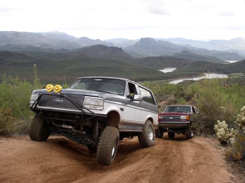 1990 Ford Bronco - Two Ford Broncos navigate a dusty trail in a scenic, mountainous desert landscape.