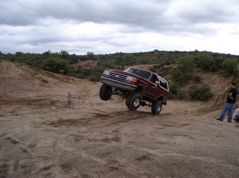 1990 Ford Bronco - A maroon Ford Bronco is captured mid-air during an off-road excursion, its lifted suspension and large tires.