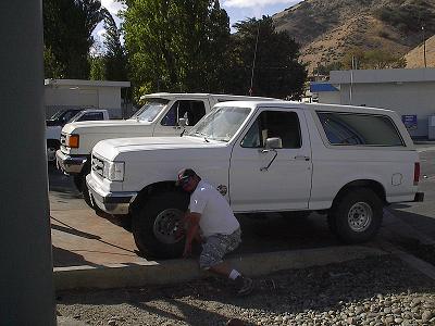 1988 Ford Bronco - A man inspects the tire of a white 1988 Ford Bronco, with another Ford truck visible behind it.