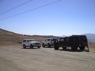 1988 Ford Bronco - Three off-road vehicles are parked on a dirt road with hills and mountains in the background.