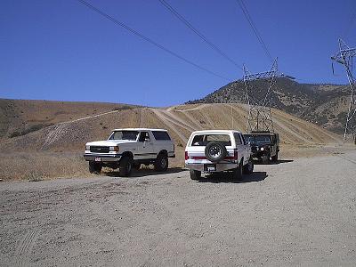 1988 Ford Bronco - Several SUVs are parked on a dirt road with power lines and a hill in the background.