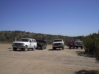 1988 Ford Bronco - Four trucks are parked in a dirt lot under a clear blue sky.