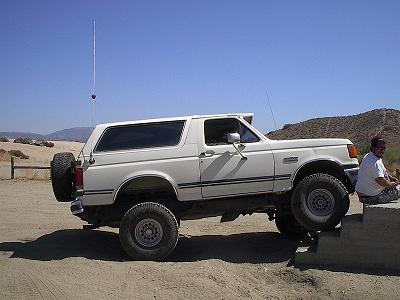 1988 Ford Bronco - A white Ford Bronco is parked on a dirt surface with a person sitting on a concrete barrier nearby.