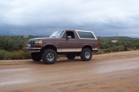 1992 Ford Bronco - Brown 1992 Ford Bronco with aftermarket wheels parked on a dirt road under a cloudy sky.